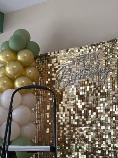 A decorative backdrop of small, gold-colored square panels is partially visible, featuring a silver "Happy Birthday" sign. Below, a balloon arrangement consists of gold, white, and green balloons. The balloons form a cluster, with varying tones of green creating depth. A black metal frame partially obstructs the lower right side of the image, adding structure to the festive decor.