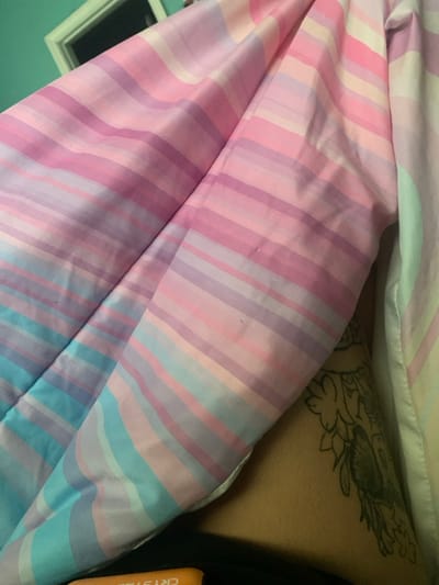 The image displays an indoor scene dominated by a pastel-striped comforter or blanket, featuring horizontal bands of pink, purple, and light blue. The comforter is folded and positioned to partially obscure the view. To the right of the comforter, a section of human skin is visible, adorned with a large, dark-inked tattoo depicting an intricate floral or organic design. In the upper left background, a teal-colored wall is partially visible, against which a white-framed object, possibly a mirror or picture frame, is mounted. At the bottom center, an orange object with partially visible white text, including the letters "CRYSTAL," rests on a dark surface. No discernible actions or interactions are depicted. The scene is situated in the city of Lancaster, United States of America.