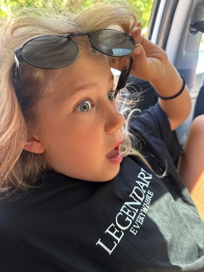 A fair-skinned child with blonde hair and green eyes is holding black sunglasses. She is lying down wearing a black t-shirt with the word "LEGENDARY" printed in white. The photograph was taken inside a vehicle, as the car doorframe is visible in the upper left corner. It is not possible to ascertain that the location is in the city of Kruče - Kërruçi, Montenegro without additional data in the image.