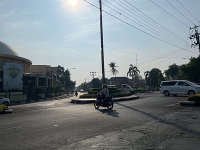 The image depicts a street scene in Bulu, Indonesia, on a bright, sunny day. The foreground shows a cracked asphalt road. A person on a motorcycle, wearing a helmet and a gray shirt, is riding through a roundabout. The motorcycle is orange and black. In the background, a white minivan is parked to the right, and a white van is visible on the left. A building with a dome and a large banner is on the left side of the road. Palm trees and other greenery are present in the midground. Electrical poles and overhead wires are prominent in the sky, which is clear with some wispy clouds. The sun is high in the sky, casting long shadows across the road.