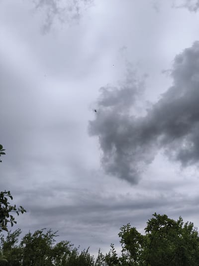 A helicopter is visible in the sky above trees in Uzunovo, Russia. The sky is cloudy and gray.