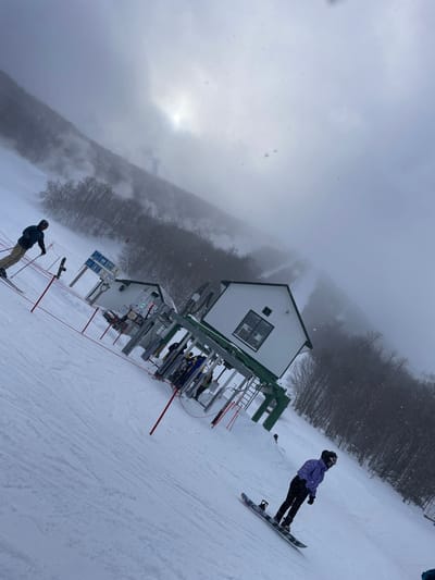 The image captures a diagonal view of a snowy ski resort, indicative of the Jay, United States location. The scene is dominated by overcast, foggy skies with light snow actively falling, creating low visibility across the distant, tree-covered slopes.
In the mid-ground, two small, white buildings with green roofs serve as ski lift stations or control points. Surrounding these structures are metal ski lift machinery, including pylons and cables, and red ropes guide queues. Several people are visible in this area, either waiting to board the lift or having just disembarked.
Two individuals are prominently featured on the snow-covered slope. On the right, a snowboarder, dressed in a purple jacket, black pants, and a helmet, descends the hill. At the bottom-left, a skier wearing a dark jacket, light pants, and a helmet, holding ski poles, is also on the snow. Ski and snowboard equipment are scattered throughout the lift area. A blue informational sign with white pictograms, typical of resort signage, is visible near one of the buildings, though specific text is unreadable. The overall atmosphere depicts a cold, active day at a ski area, with enthusiasts enjoying winter sports despite the foggy and snowy conditions.