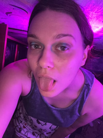 The image shows a close-up, selfie-style shot of a person with their tongue sticking out. The lighting is predominantly purple, suggesting an artificial light source, possibly a colored LED bulb or a filter. The person is wearing a dark-colored tank top over a patterned garment, possibly pajamas, with cartoon-like illustrations. A gold hoop is visible in their nose. The background is blurry and appears to be indoors, with some wooden furniture and a ceiling or wall illuminated by the purple light. There is no indication of the city of Bishkek, Kyrgyzstan, in the image.