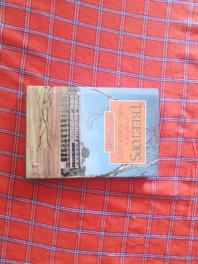 The image shows a paperback book lying on a red and blue checkered cloth. The book's cover features a photograph of a building in the background and a tree in the foreground. The book's title is "Treetops: Story of a World Famous Hotel" by R.J. Pricket.