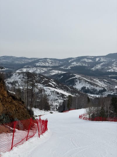 The image depicts a wide, groomed ski slope within the city of Novoabzakovo, Russia, under an overcast, light gray sky. The foreground features a snow-covered ski run, exhibiting parallel grooming marks, which curves gently towards the right. Red safety netting fences delineate both edges of the slope. To the left, a steep, reddish-brown rocky embankment with scattered bare trees borders the run. To the right, the slope is bordered by a snow-covered area containing a mix of bare deciduous and evergreen trees. In the midground, the ski slope descends into a valley. The background presents an expansive view of multiple layers of snow-covered mountains and hills, characterized by dark, sparse tree cover, extending to the horizon. No individuals or active subjects are present in the scene.