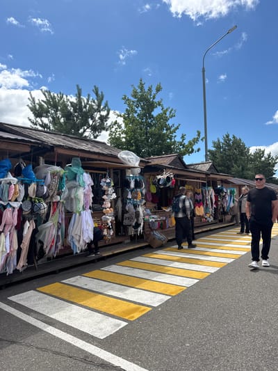 The image depicts a street scene in Privolzhsky, Russia, featuring a market stall laden with various goods. Two people are visible in the foreground, standing near a pedestrian crossing marked with yellow and white stripes on an asphalt road. The stall itself is a wooden structure overflowing with merchandise, including hats, clothing items, and small plush toys. The items are displayed on shelves and hanging racks, creating a vibrant and somewhat cluttered visual.

In the background, trees with lush green foliage are prominent against a bright blue sky dotted with white clouds. A street lamp with a modern design arches over the scene, suggesting it's daytime. The overall atmosphere appears to be that of a bustling outdoor market on a clear, sunny day. The presence of the pedestrian crossing and the open-air market setting implies a public space with pedestrian traffic. No visible text is discernible in the image. The scene conveys a sense of everyday commerce and community activity.