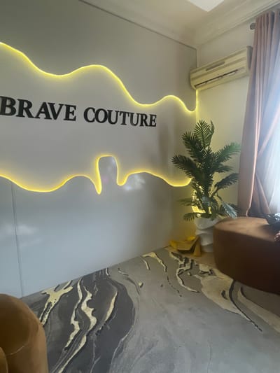 The image, oriented sideways, captures an interior scene, likely a commercial establishment such as a boutique or salon. A dominant feature is a white wall adorned with a large, wavy, abstract light fixture that glows with a warm yellow hue. Affixed to this wall is prominent black text that reads "BRAVE COUTURE."
To the right of the wall, a verdant, palm-like plant, possibly artificial, is housed in a white pot. Portions of a brown upholstered piece of furniture, appearing to be an armchair or sofa, are visible at the top and bottom right, suggesting a seating area. The floor is covered by a patterned carpet, predominantly grey, with abstract swirls and lines in lighter grey, gold, and black, resembling a marble or geological formation.
Near the ceiling, an off-white wall-mounted unit, likely an air conditioner or a similar utility fixture, is present. Above the furniture, wooden blinds or slats, partially obscured by sheer curtains, hint at a window or an opening. The lighting in the room is soft and ambient, largely provided by the glowing wall fixture, contributing to a welcoming atmosphere. No individuals are visible, and no specific activity is depicted. The presence of "BRAVE COUTURE" indicates a fashion-related business. The location is Dushepe, Nigeria.