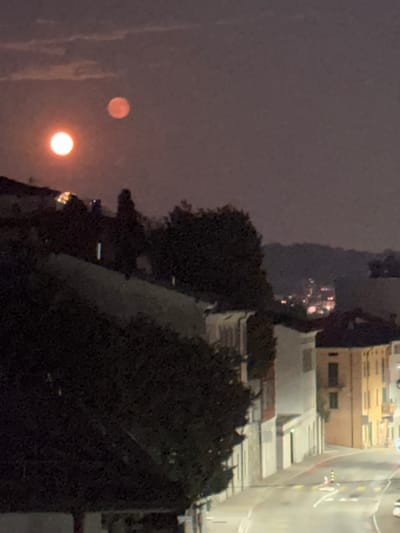 The image, taken at night in Balerna, Switzerland, shows a view of a street and buildings with two luminous celestial bodies in the sky. The larger, brighter body, appearing to be the moon, is a white-orange orb in the upper left portion of the frame. To its upper right, a smaller, reddish orb, also seemingly a moon or a distinct celestial event, is visible. The foreground features a dark, leafy tree on the left and a sloped embankment. Further back, a street curves through a built-up area with buildings of varying heights and colors, some with visible windows and balconies. Streetlights and other artificial lights illuminate parts of the scene, reflecting on the wet-looking road surface. The background shows distant hills or mountains silhouetted against the night sky. No people are discernible in the image.