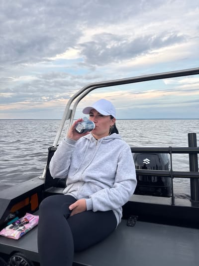 A medium shot captures a woman in a gray zip-up hoodie and black leggings, wearing a white baseball cap, sitting on the deck of a boat. She is holding a clear plastic bottle with a gray lid and dark liquid to her mouth, as if drinking. The boat is on a large body of water, likely a lake, with a cloudy sky overhead. In the background, the horizon is visible, separating the water from the sky. A black boat motor with a white Suzuki logo is mounted to the rear of the boat, partially obscured by black railing. To the left of the woman, on the deck, is a pink package of wet wipes, along with a dark bottle and a green object. The overall scene suggests a day trip on the water. The location is identified as Kostino, Russia.