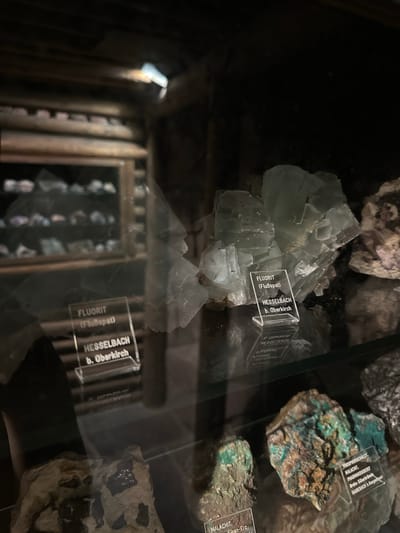 The image shows a display case in what appears to be a museum or a private collection. The focus is on various mineral specimens. In the foreground, there are several mineral samples with labels. One prominent specimen is a large, pale green crystal formation, likely fluorite, as indicated by a label that reads "FLUORIT (Flußspat) HESSELBACH b. Oberkirch". The label is made of clear acrylic and is standing upright. Reflected in the glass of the display case, another similar label can be seen, suggesting a mirrored arrangement or a second display.
Further back, other mineral samples are visible on shelves, some appearing smaller and in various colors. The display is dimly lit, with a light source illuminating the specimens from above. The background suggests a wooden structure, possibly part of the museum's architecture.
The mention of "Hesselbach b. Oberkirch" on the label points to a specific geographical location within Germany, a region known for mineral deposits. While the image itself doesn't explicitly show Schonach im Schwarzwald, the context of mineral displays and the origin of specimens from areas like Oberkirch are consistent with a mineralogical exhibit that might be found in or related to the Black Forest region.