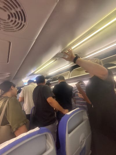 The image shows a group of people standing in the aisle of an airplane, likely boarding or disembarking. A woman in the foreground on the right, wearing a black top and a watch, is reaching up towards the overhead compartment. Other passengers are visible in the background, some standing and some seated in blue and white airline seats. The interior of the plane is lit by overhead fluorescent lights. There is a circular air vent visible on the left side of the ceiling. The image is taken from a low angle, looking upwards.