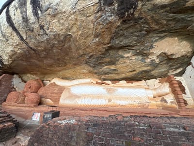 The image shows a large, partially preserved reclining Buddha statue located in Sigiriya, Sri Lanka. The statue lies beneath a rocky overhang that shelters it from the elements. The statue's head rests on a stacked-brick pillow structure, and its body is mostly white with layered grooves on its robes. The statue is positioned on a low brick platform that is also partially ruined. In the foreground, there is a dark, mossy brick structure. To the left is a black display case with a sign in front. A thin wire is strung along the front of the statue.