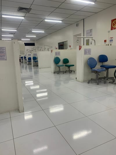 This image displays a brightly illuminated, unoccupied dental practical laboratory or simulation room located in Lauro De Freitas, Brazil. The environment features a uniform white aesthetic, with a glossy white tiled floor reflecting numerous rectangular overhead fluorescent lights set within a white grid ceiling. The room is structured by rows of white, waist-high partitions, creating individual workstations. Each visible workstation is equipped with an overhead adjustable dental examination light, a water spigot, and often two rolling, adjustable chairs with casters. In the foreground right, two light blue and two darker blue chairs are present. In the mid-ground left, two teal-green chairs are visible. Red numerical indicators, such as "22," are affixed to the partitions alongside printed informational signs. A red circular warning sign is mounted on the far right wall. No individuals are present, and no actions or interactions are occurring within the scene.