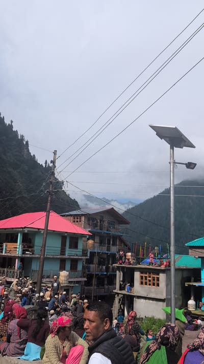 The image depicts a crowded scene in the village of Malana, India, situated amidst mountainous terrain. A densely packed group of people, many wearing traditional caps, are gathered in an outdoor area. Buildings with colorful roofs, predominantly red and teal, line the background, with some appearing to be multi-story structures. Several individuals are seated, while others are standing, suggesting a communal gathering or event. Electrical wires crisscross the sky, and a solar-powered street lamp stands prominently on the right side of the frame. The environment is characterized by a hazy, overcast sky and the imposing presence of lush green mountains in the distance. The overall impression is one of a vibrant community life set against a rugged natural landscape.