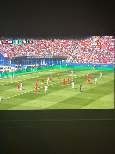 This is a photograph taken from a screen, likely a television, showing a football match. The scene is a brightly lit football stadium with a vibrant green pitch. In the foreground, there are several players in red and white kits, actively engaged in a game. The stands are packed with spectators, predominantly dressed in red and white, creating a colorful and energetic atmosphere.

Along the side of the pitch, a green banner displays various text and logos. Visible text includes "FIFA WORLD CUP 2026", "NEXTSTARTSNOW", and "HYUNDAI", suggesting this is a promotional display for the upcoming FIFA World Cup. Another section of the screen shows a scoreboard or information overlay with "NOR 1 2 FRA" and "НА ЖИВО 1 200 1", indicating a match between Norway (NOR) and France (FRA) with a score of 1-2, and "Live" in Cyrillic. There is also a reference to "DUCE".

The orientation of the image suggests it was taken with the camera held vertically, or the screen is rotated. The lighting appears to be daylight, as indicated by the shadows cast by the players on the field. The overall impression is one of an exciting sporting event within a well-attended stadium, with clear branding from FIFA and its sponsors. The setting is a football pitch, and the event is a live match.