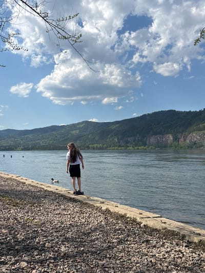 A young woman stands on a concrete embankment alongside a river in Овсянка, Russia. She wears a white shirt and black skirt. The river is wide and calm, with a mountain visible in the background. The sky is blue with scattered clouds. To the right of the embankment is an area covered in small stones. A duck floats near the edge of the embankment.