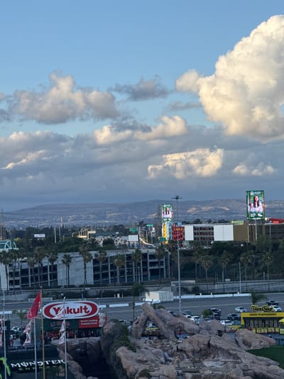 The image is taken in Anaheim, United States. The sky is divided into light blue and dark gray clouds. There are mountains visible in the distance. There are two billboards displaying the logo of California University, one on top of the other. There is a lot of building construction throughout the landscape, with many palm trees. There is a Topo Chico stand, several parking lots, and a Yakult billboard visible. There are several flags.