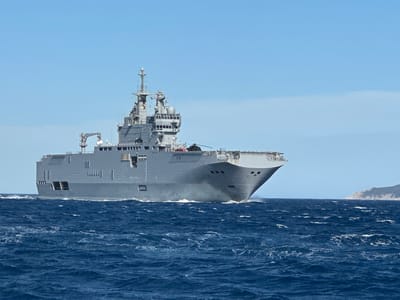 A large, grey naval vessel is sailing through choppy, dark blue ocean waters under a clear, light blue sky. The ship has a prominent superstructure with multiple levels and a tall mast with antennas. A helicopter can be seen on the deck of the ship towards the stern. In the distance, on the right side of the frame, a landmass, likely an island or coastline with vegetation, is visible. The ship is creating a wake as it moves through the water. The overall impression is one of a naval exercise or deployment in an open-water environment. This scene is not located in the city of Bormes-les-Mimosas, France; it depicts a maritime scene.