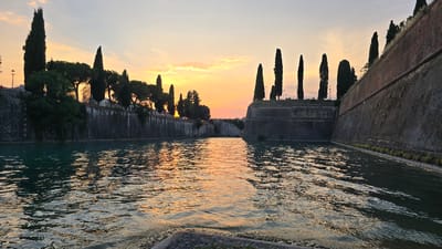 The image depicts a canal in Peschiera del Garda, Italy, at sunset. The water reflects the orange and yellow hues of the sky, creating a shimmering effect. Tall cypress trees line the stone walls of the fortress, casting long shadows. The fortress walls are made of rough stone and bricks, with some vegetation growing on them. The sky is a soft gradient of orange, pink, and yellow, with a few wispy clouds. The scene is serene and picturesque, capturing the beauty of the Italian landscape at dusk.