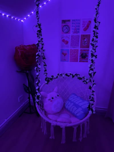 The image depicts a dimly lit room, bathed in purple light from LED strip lights along the ceiling. A macrame swing chair, adorned with a vine of white artificial flowers, is centrally positioned. Inside the chair, two plush pillows are visible: one is a light pink bunny with black eyes and stitched whiskers, and the other is a blue pillow with white text that appears to say "BIG BLUES." Next to the swing chair, a large, red balloon shaped like a rose stands tall. The wall behind the chair features a grid of nine small posters, each with a different design and text. These posters include graphics of a globe with text "MIRROR BALL", multiple "PARIS" text prints, a red flip phone with the text "DON'T CALL ME", two red dice with text "DEVILS ROLL THE DICE", a pink pattern with "FLOWER MARKET", a glass with a pink drink and text "MOJITO", a silhouette of buildings with text "TOKYO", and two sets of text "ROME". Further posters include text "disco disco disco". The floor is a dark wood laminate. No people are present in the image. The location is identified as Obninsk, Russia.