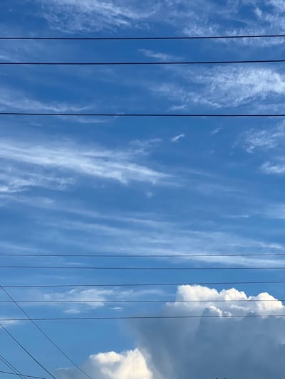 The image is an outdoor, eye-level shot of a bright blue sky filled with wispy white clouds. Several dark, horizontal power lines stretch across the frame, intersecting the sky and clouds. In the lower right quadrant of the image, large, fluffy white cumulus clouds are visible, suggesting a sunny day. There are no people, animals, or buildings discernible in the image. The scene appears to be a typical sky view, possibly from a residential or semi-rural area. The location is identified as Pyogozino, Russia.