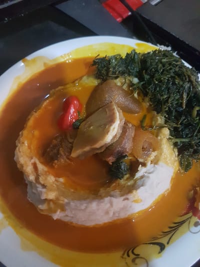 The image shows a close-up, overhead view of a plate of food. The food appears to be a starchy base, possibly fufu or mashed yams, covered in a rich, orange-colored stew. The stew contains visible chunks of meat, some with a gelatinous texture, and a small red chili pepper. To the right of the stewed portion is a pile of dark green, leafy vegetables, likely a type of spinach or indigenous greens. The plate itself is white with a yellow border and decorative black patterns. The food is presented on a dark surface, possibly a table or countertop, with a blurred dark background that suggests an indoor setting. No people or specific landmarks are visible, making it impossible to determine the exact location within Yaoundé, Cameroon. The lighting is artificial, casting a warm glow on the food.