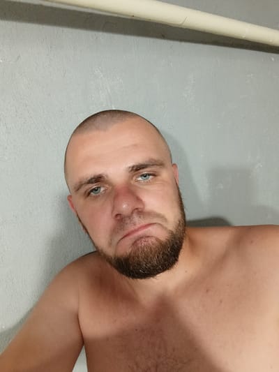 A man with a shaved head and beard poses for a selfie in front of a grey, textured wall. A white pipe runs horizontally across the top of the frame. The man's expression is downturned, with his mouth slightly open and his eyes looking towards the camera. His bare shoulders are visible. The lighting is artificial and casts a shadow to the right of the man. The image appears to be taken indoors, possibly in a residential setting.