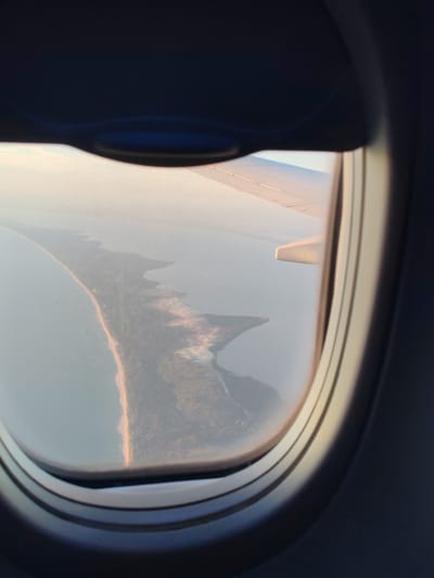 The image captures an aerial view from an airplane window, looking out over a coastline. The primary subject is a long, narrow peninsula or strip of land extending into the ocean. The land appears to be covered in vegetation and shows signs of a fire, with areas of burnt landscape visible. A sandy beach borders the peninsula on one side, meeting the calm, pale blue water. The overall scene is bathed in the soft, warm light of either sunrise or sunset, casting a serene glow on the landscape and the aircraft's wing, which is partially visible in the upper right and left sections of the frame. The time of day is suggested by the warm, low-angled light. The weather appears clear and calm. The view from the window, framed by the dark interior of the airplane cabin, creates a sense of travel and observation. There is no visible text in the image.