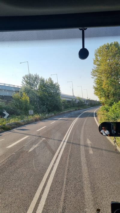 The image captures a view from inside a vehicle, looking out onto a two-lane asphalt road with clear white lane markings. The road curves gently to the left. To the left of the road, there is lush green vegetation and trees, with a concrete overpass visible in the distance. Several streetlights are spaced along the side of the road. In the foreground on the left, a dark circular object, possibly a dashcam or GPS device, is mounted on the windshield.

The sky is a clear blue, suggesting a sunny day. The time of day appears to be morning or late afternoon, as evidenced by the angle of the sunlight creating shadows on the road. A side-view mirror is visible in the upper right corner of the image, reflecting a portion of the interior of the vehicle and a person wearing a white shirt and a light-colored hat. The overall scene is tranquil and suggests a journey through a semi-rural or suburban area. There is no visible text in the image.