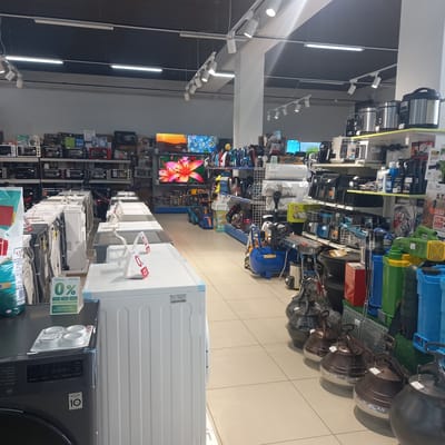 The image depicts the interior of a retail store, likely an appliance or electronics store, located in Briceni, Moldova. The scene is brightly lit with overhead fluorescent and track lighting. In the foreground, a row of white washing machines is visible, with a dark gray washing machine on the left. Several of these appliances have "0%" financing offers displayed on white stands.

The middle ground shows a variety of appliances and home goods arranged on shelves and displays. This includes pressure cookers, fans, and other kitchen appliances. In the background, shelves are stocked with boxes of electronics and appliances. Television screens are also visible, displaying colorful images. The store has tiled flooring and a modern, organized layout. There are no people visible in the image. The overall atmosphere is that of a well-stocked retail environment.