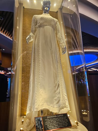 This is an interior shot taken at the Seminole Hard Rock Hotel & Casino in Hollywood, Seminole Reservation, USA. It features a mannequin dressed in a white dress, presumed to be a vintage 1970s wedding gown, encased in a glass display. The display includes a plaque that identifies the dress as being worn by Jennifer Lopez in the wedding scene of the 2007 film 'El Cantante'. The background of the display is a textured golden material, and the room is somewhat dimly lit. Reflections in the glass casing suggest other elements of the interior, including ceiling lights and possibly the layout of the casino area. There are no people clearly visible in the immediate foreground or directly interacting with the display.