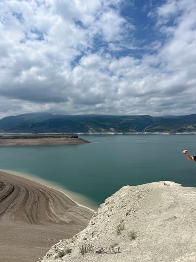 The image depicts a view from a high vantage point overlooking the Chirkey Reservoir in Chirkey, Russia. The reservoir fills most of the frame, with the water reflecting the cloudy sky above. A narrow strip of land, exposed due to a possible drop in water level, is visible along the reservoir's edge. The coastline is layered and textured. On the left, the mountainous terrain rises steeply. Part of a person's arm is visible at the top edge of the image. The edge of the cliff or rock formation that the image is taken from is visible to the right.