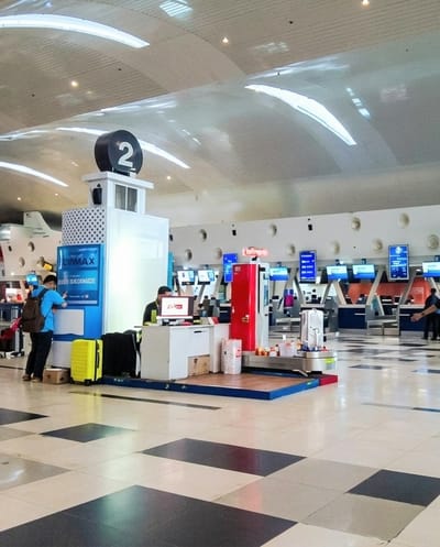 The image captures an indoor scene within an airport terminal, likely a check-in or baggage drop-off area in Sidourip, Indonesia. The spacious room features a high, white, curved ceiling with embedded strip lighting and spotlights, illuminating the space brightly. The floor is tiled in a distinct checkerboard pattern of dark and light squares.
In the mid-ground, a central kiosk is prominent, topped with a black cylindrical sign displaying the number "2." One side of this kiosk features a blue sign with "JETMAX" and "BORDER GUARANTEE" visible text. To its left, a person wearing a blue shirt and a brown backpack is interacting with a screen at a counter, suggesting self-service check-in or baggage processing. A bright yellow suitcase rests on the floor beside them.
Adjacent to the central kiosk, a person in a dark shirt is seated at a white desk, focused on a computer monitor, appearing to be an airport staff member. Next to this desk is a tall, red kiosk or machine with an orange display, and the word "DELIVERY" partially visible above it. A small, white, wheeled suitcase is positioned on a baggage carousel-like structure near this red kiosk, alongside rolls of white wrapping material.
In the background, numerous check-in counters are visible, characterized by blue and white structures and screens, indicating a larger check-in facility. The walls behind the counters feature circular indentations. The overall atmosphere is calm, with individuals engaged in routine travel activities, suggesting daytime hours due to the bright, artificial lighting. No specific emotions are discernible, but the scene appears functional and orderly.