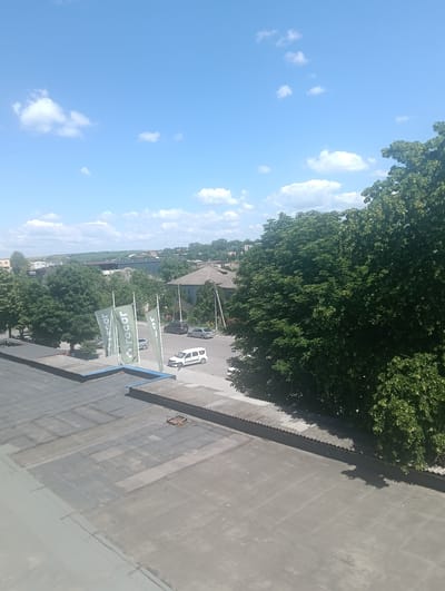 This image depicts a street scene in Florești, Moldova, on a clear, sunny day. In the foreground, a flat, grey rooftop is visible, marked with a grid pattern of darker grey squares. Beyond the rooftop, a street runs horizontally across the frame. On the street, several vehicles are present: a white van, a white sedan, and a dark-colored sedan. Three tall, green flags with white logos are planted along the side of the street, featuring the text "rocar" repeated. Lush green trees dominate the right side of the frame, with their dense foliage casting shadows. In the background, a town or city landscape unfolds, characterized by low-rise buildings, more trees, and rolling hills covered in green fields stretching towards the horizon under a bright blue sky dotted with white clouds. The overall impression is one of a typical day in a provincial town.