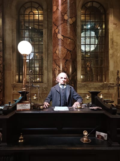 The image shows a scene likely from a movie set or museum exhibit replicating the Gringotts Wizarding Bank. A goblin bank teller, wearing a grey pinstripe suit and a stern expression, sits behind a dark wooden desk. On the desk are several items including a balance scale, inkwell, papers, and small brass bells. A sign on the desk identifies the character as a "GOBLIN BANK TELLER" and mentions the actor. Behind the desk are large windows with ornate frames, and between them a tall, patterned pillar. The location, according to the data provided, is in Three Rivers, United Kingdom. The background suggests a grand and magical setting typical of the Harry Potter series.