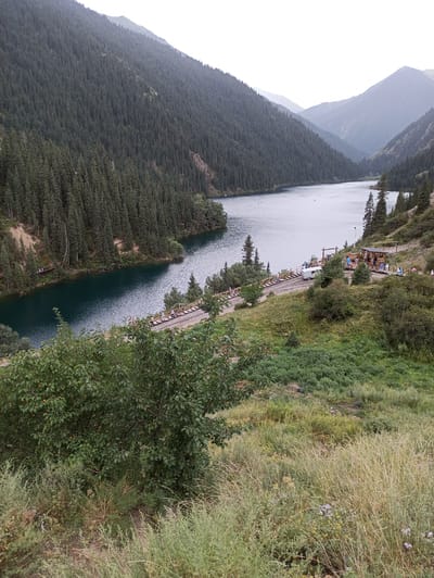 The image depicts a panoramic view of the Kolsai Lakes in the Tien Shan mountains, near the village of Kurmeti, Kazakhstan. A serene, deep blue lake is nestled between towering, heavily forested mountains. The slopes are densely covered with evergreen trees, creating a lush, natural landscape. Along the shoreline, a wooden walkway or path is visible, with several people strolling and enjoying the scenery. A small wooden structure, possibly a cafe or rest area, is situated near the water's edge, with a white caravan parked nearby. The foreground features a slope covered with dry grass and shrubs, suggesting a somewhat arid or transitional season. The overall atmosphere is one of natural beauty and tranquility, with signs of human presence integrated into the environment.