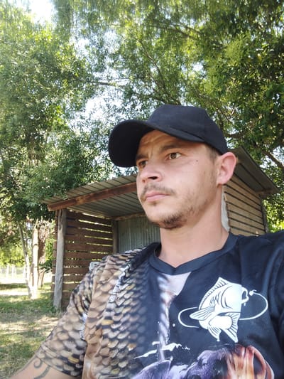 A man wearing a black baseball cap and a shirt with a fish design on the chest and what appears to be scale patterns on the shoulder is the subject of this outdoor selfie. He is clean-shaven and has a light complexion. Behind him is a small, wooden shed with a corrugated metal roof and slats for walls, set against a backdrop of trees and greenery. The location of Sant'Andrea del Pizzone, Italy cannot be confirmed.