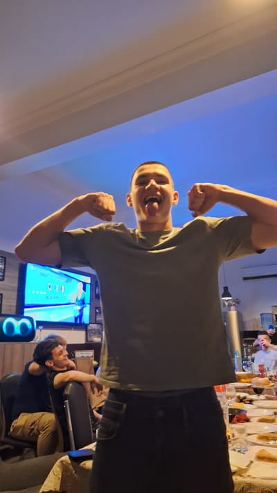 A tilted, low-angle indoor shot. A young man with a shaved head is facing the camera, sticking his tongue out and flexing both arms with clenched fists. He wears a gray t-shirt and dark pants. In the background, a TV screen displays a game with brightly colored graphics. To the right, a man with curly hair is seated in a chair, his head resting on his arm. A table laden with plates and food is visible in the far background. The location is likely a private residence, but the specific details needed to determine it being Gorski Izvor, Bulgaria, are unavailable.