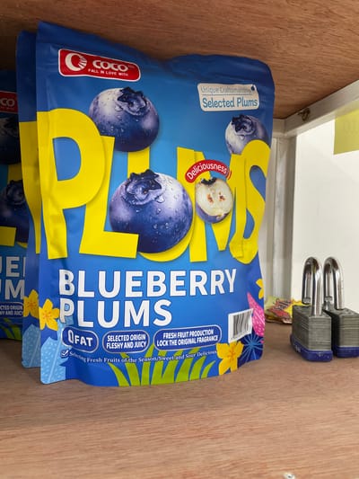 The image depicts the interior of what appears to be a storage area or a shelf in a commercial establishment, possibly a warehouse or a shop, located in San Jose del Monte, Philippines. The dominant subject is a large, bright blue bag of "BLUEBERRY PLUMS" manufactured by "COCO." The bag features prominent illustrations of blueberries and abstract floral patterns in yellow and blue. Text on the bag highlights attributes such as "Unique Craftsmanship," "Selected Plums," "Deliciousness," "0 FAT," "Selected Origin Fleshy and Juicy," and "Fresh Fruit Production Lock the Original Fragrance." There are visible expiration dates on the bag. To the right of the bag, two silver padlocks with blue bases are placed on a shelf, suggesting a security measure. The background reveals wooden shelving and a white wall. The lighting suggests an indoor, possibly artificial, light source.