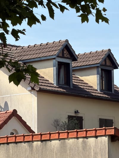 A sunny outdoor shot captures the upper portion of a light-yellow building in Rungis, France, under a clear blue sky. Dark green leaves from a tree hang into the frame from the top left. The building features a brown tiled roof with two dormer windows. The dormer windows are framed with grey metal and have dark windowpanes. One dormer window has sheer white curtains visible. Below the main roof, a section of a lighter-colored wall is visible, leading down to a low, textured grey wall. Along the top of the grey wall, there is a row of red tiled elements.