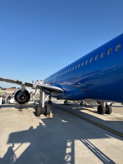 The image depicts a blue passenger airplane parked on a concrete tarmac, presumably at an airport. The plane's body is visible, showing a row of passenger windows. Landing gear is visible underneath the fuselage. People are boarding via an external staircase attached to the plane. The setting appears to be outdoors on a sunny day, given the clear blue sky. The architecture of nearby buildings suggests a European location. Although it is impossible to verify from the images, the photo is purportedly taken at Peschiera Borromeo, Italy.