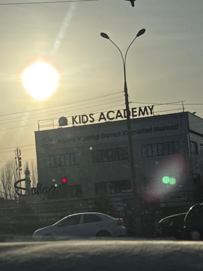 A building marked "KIDS ACADEMY" stands beside a street. The sign is partially visible. In the background, a bright sun shines beside a street lamp. Part of a vehicle, likely a car, is visible on the right edge of the image. Some power lines and another building can be seen in the distance.
