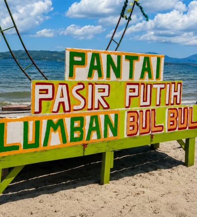 A brightly painted, multi-tiered sign in white, green, and orange reads "PANTAI PASIR PUTIH" on the top tier, and "LUMBAN BUL BUL" on the bottom tier. The sign is situated on a sandy beach with the ocean in the background. The water is blue and calm, meeting a hazy horizon line with distant hills. The sky is partly cloudy with white cumulus clouds against a bright blue sky. The sign is supported by a green wooden structure. The overall scene depicts a tropical beach location. The location is identified as Lumban Bul Bul, Indonesia, a village known for its beaches.