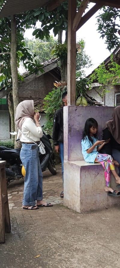 The image shows a scene in Cimaung, Indonesia, located at latitude -7.074 and longitude 107.547. Three individuals are present. Two women and a man are standing in front of a concrete structure. The women are both wearing sandals. One woman is wearing a light-colored sweater over a white t-shirt and blue jeans. She is looking directly at the camera. The other woman is wearing a blue t-shirt and colorful pants. She is looking at a phone in her hand. The man is wearing a black jacket. He is looking down at the ground, with his hands in his pockets. The concrete structure has a bench on top of it with a purple wall.  The scene is outdoors, in a residential area. There are trees in the background and a motorcycle parked in front of the structure.