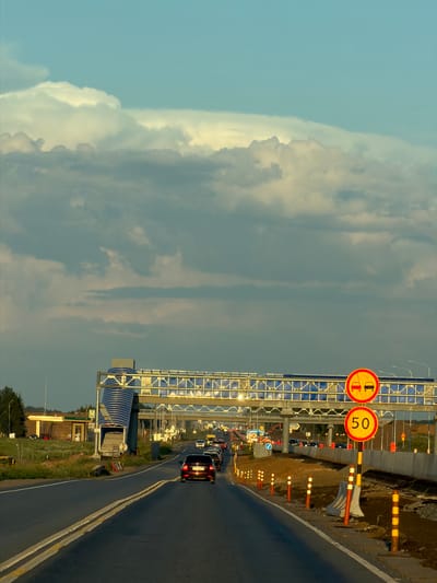 The image captures a highway scene in Derzhavino, Russia, with a car driving on the road. The sky is filled with dramatic clouds, hinting at an approaching storm or sunset. Traffic signs indicating a speed limit of 50 km/h and a prohibition on overtaking are prominently displayed on the right side of the road. Construction barriers and cones suggest ongoing roadwork. A pedestrian overpass spans the highway in the background, and a gas station is visible on the left side of the road. The overall atmosphere is one of travel and potential weather changes.