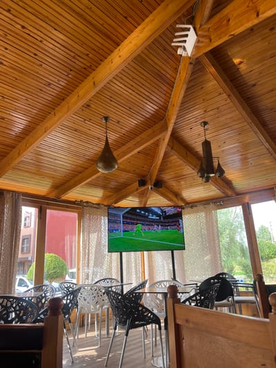 The image depicts the interior of a rustic, wooden-beamed structure, likely a cafe or restaurant, with a view of an outdoor area. The ceiling is constructed from polished wooden planks and beams, with several decorative light fixtures hanging from it. One fixture, a multi-bulb lamp with an ornate, golden shade, is prominently visible. Another white, geometric object, possibly a Wi-Fi router or a decorative element, is mounted on a beam.
A flat-screen television is mounted on a wall and displays a soccer game, with players in red and blue uniforms on a green field. In front of the television are several chairs, a mix of black and white, with intricate, modern designs. The chairs are arranged around tables, though the tables themselves are mostly out of frame.
Large windows run along one side of the structure, offering a glimpse of the outside. Through the windows, trees and greenery are visible, suggesting a pleasant, perhaps sunny, day. Sheer, patterned curtains soften the light coming through the windows. The lower portion of the windows reveals more of the outdoor environment, including a red-tiled building and a white vehicle. The overall atmosphere is casual and relaxed, suggesting a place where patrons might gather to watch sports or enjoy a meal.
No discernible emotions are conveyed, and the time of day appears to be daytime, given the natural light. The location context of El Hajeb, Morocco, does not provide specific visual cues within the image itself, but it informs the setting of the scene. No visible text is present in the image.