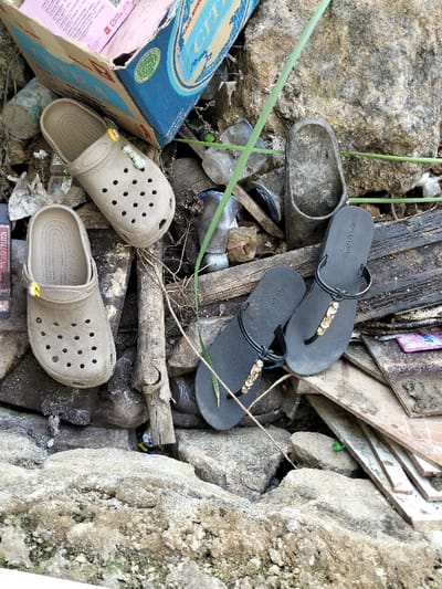 An outdoor scene showcases discarded items amongst rocks and debris. A pair of beige Crocs with decorative charms sits near a blue and beige cardboard box. A pair of black flip-flops adorned with gold accents lies beside a worn-out clog. Plastic bottles, scraps of wood, and other unidentifiable garbage are scattered throughout the scene, partially obscured by the rocks. A single blade of grass pierces the clutter.
