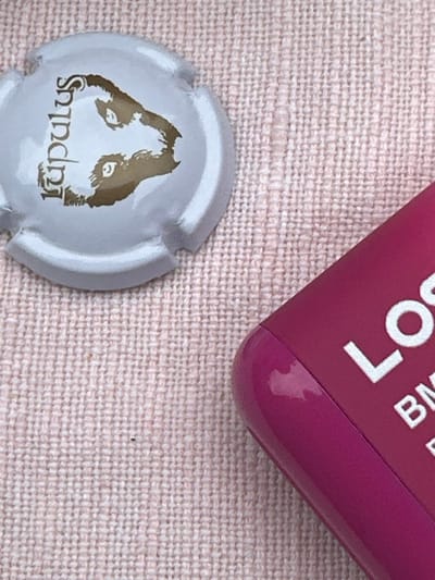 The image shows a white champagne cork with the word "Lupulus" written on it in gold lettering. It also depicts a golden dog head with black eyes. The cork is lying on a pink cloth with a woven texture. A partially visible fuchsia-colored rectangular box with a shiny surface is in the bottom right of the image. The box has the word "LO" printed in white on it. The location of the scene is likely a home or an outdoor setting in Ixelles, Belgium, as the coordinates provided are for the municipality of Ixelles.