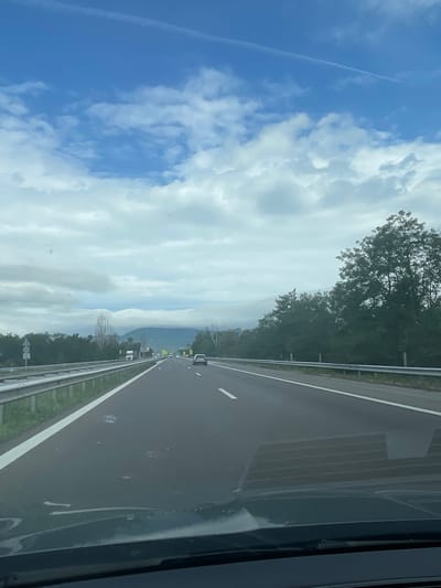 The image shows a view from inside a vehicle traveling on a highway, likely in or near Tserovo, Bulgaria. The road is paved, with clear white lane markings. A metal guardrail runs along the left side of the highway, separating it from a grassy shoulder. On the right side of the highway, dense green trees line the road. In the distance, a mountain range is visible under a cloudy sky. Several vehicles, including cars and trucks, are present on the highway. The sky is partly cloudy with a visible contrail. The reflection of the car's dashboard and windshield wipers is visible on the lower part of the image.