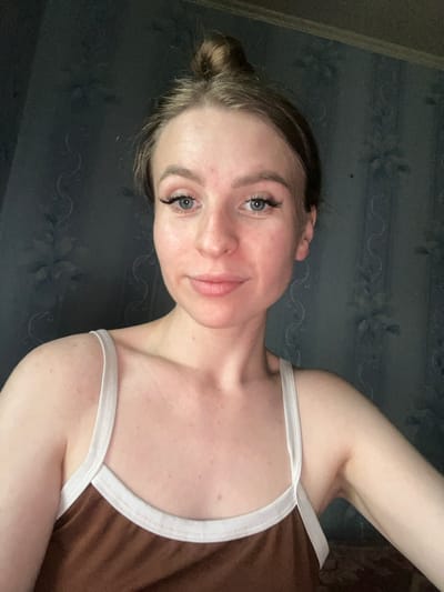 A young woman with her hair pulled up in a bun is looking at the camera. She is wearing a brown tank top with white trim. The background is a dark blue patterned wallpaper. The image appears to be taken indoors. The woman has makeup on, including eyeliner and mascara, and her skin has some redness and freckles. She has a neutral expression on her face. The lighting is soft and somewhat dim.